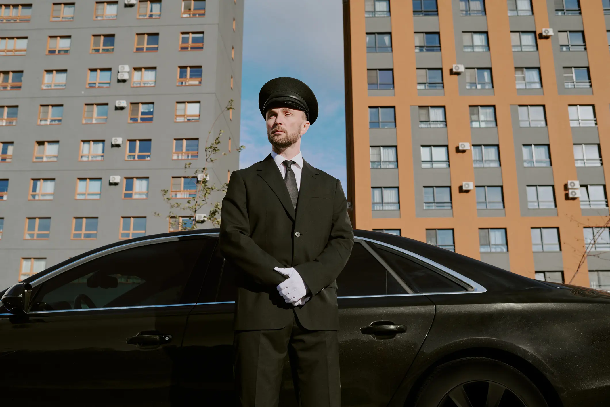 Chauffeur in cap and white gloves waiting beside a black luxury sedan in a city setting